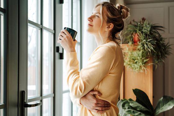 Bleib gut informiert zu aktuellen Entwicklungen auf dem Energiemarkt mit Yello Eine junge Frau steht mit einem Kaffeebecher am Fenster in ihrer Wohnung und schaut hinaus