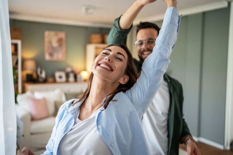 Einfach doppelt gute Laune mit Prämie auf deinen Energievertrag Ein junges, fröhliches Paar tanzt zusammen zuhause im Wohnzimmer