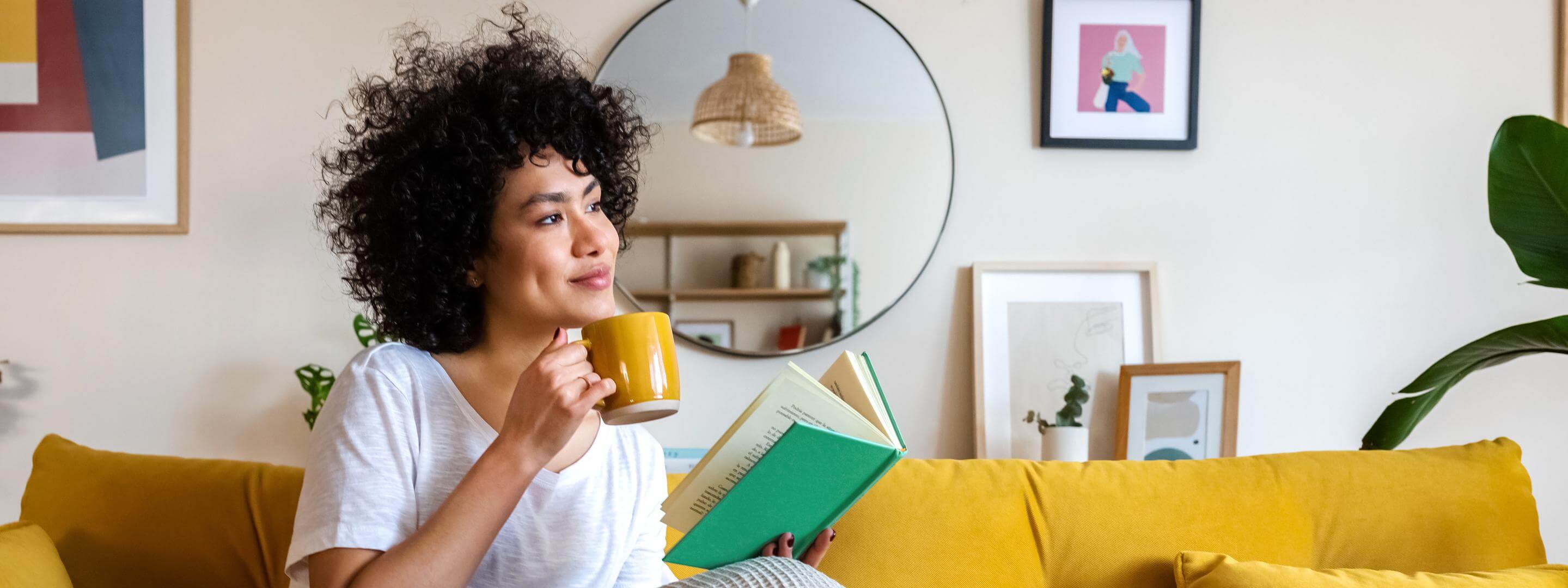 Junge Frau sitzt auf dem Sofa und liest ein Buch mit einer Tasse in der Hand.