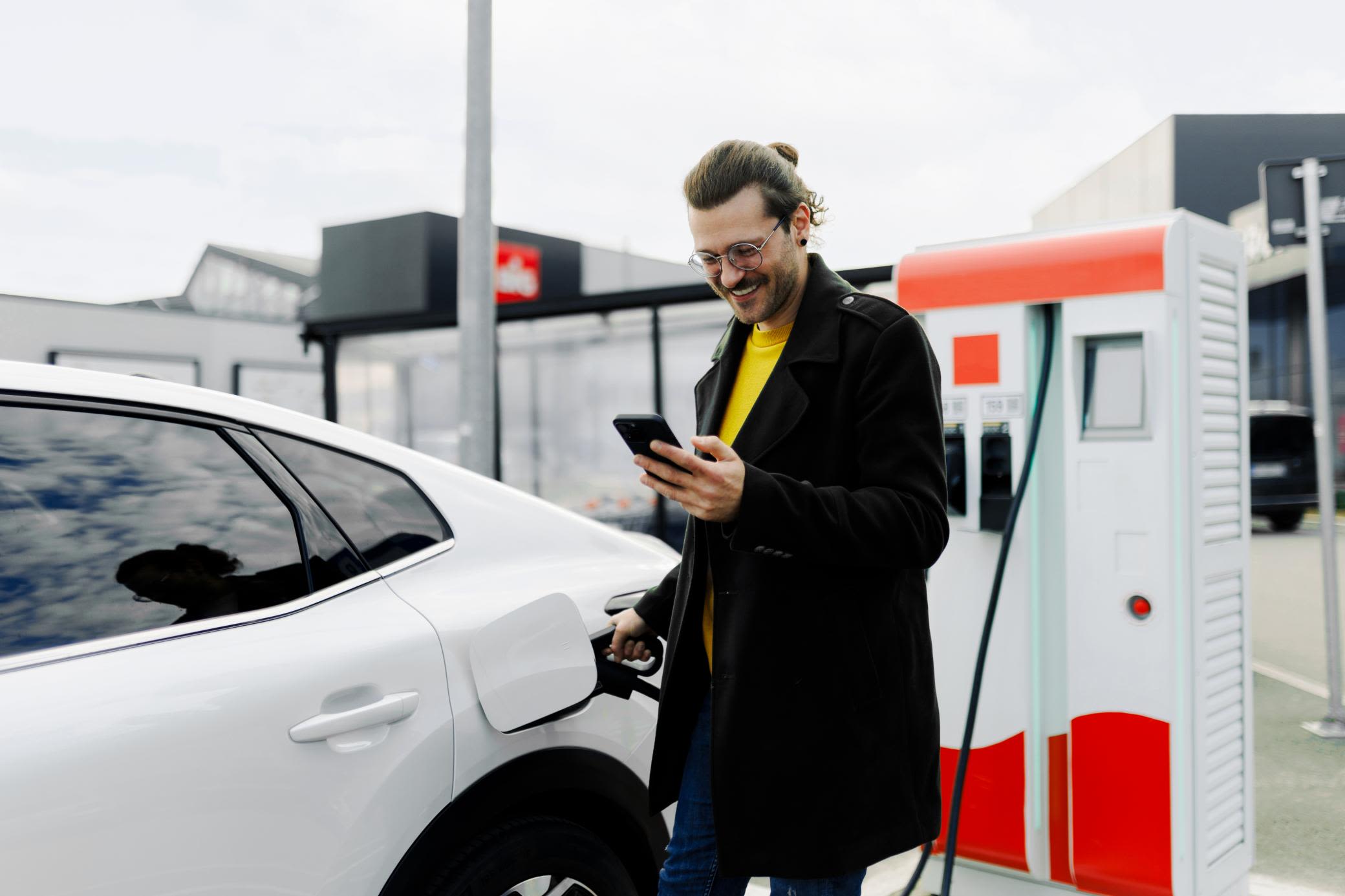 Unterwegs laden geht per App einfach und unkompliziert Ein lächelnder, junger Mann mit Smartphone lädt ein E-Auto an der öffentlicher Ladesäule