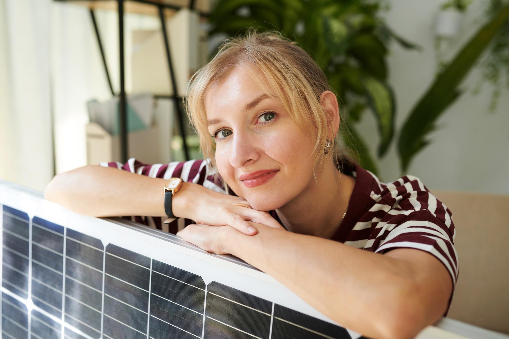 Junge Frau mit blonden Haaren lehnt zufrieden über ihrem Solarpanel in ihrer Wohnung