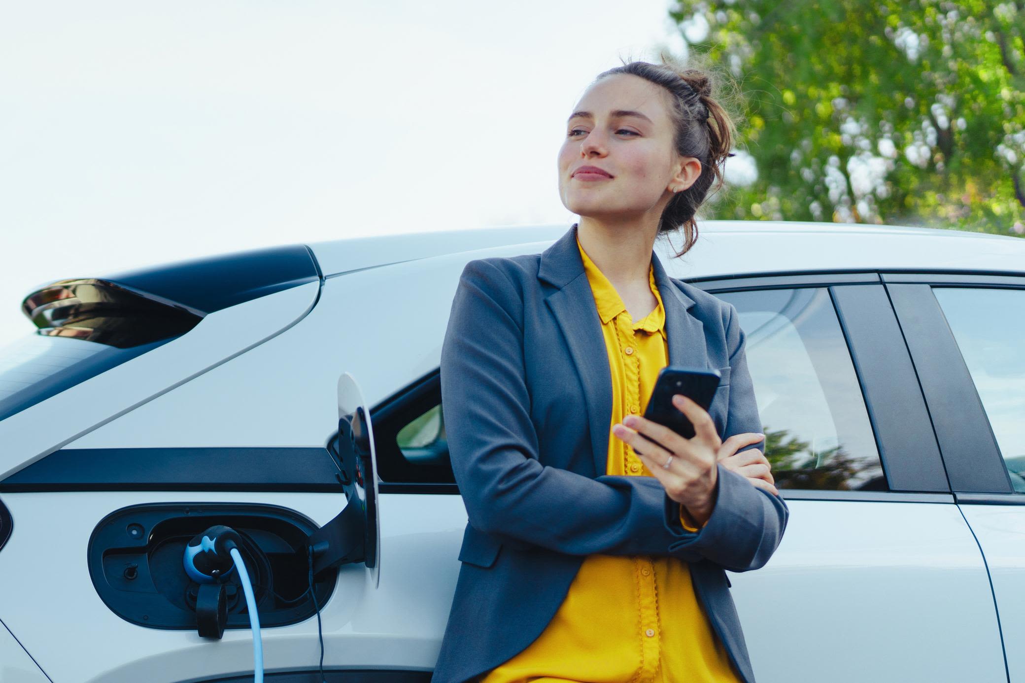 Mit Yello Autostrom zuhause und unterwegs laden Junge Frau mit gelber Bluse und Smartphone in der Hand entspannt beim Laden ihres Elektroautos