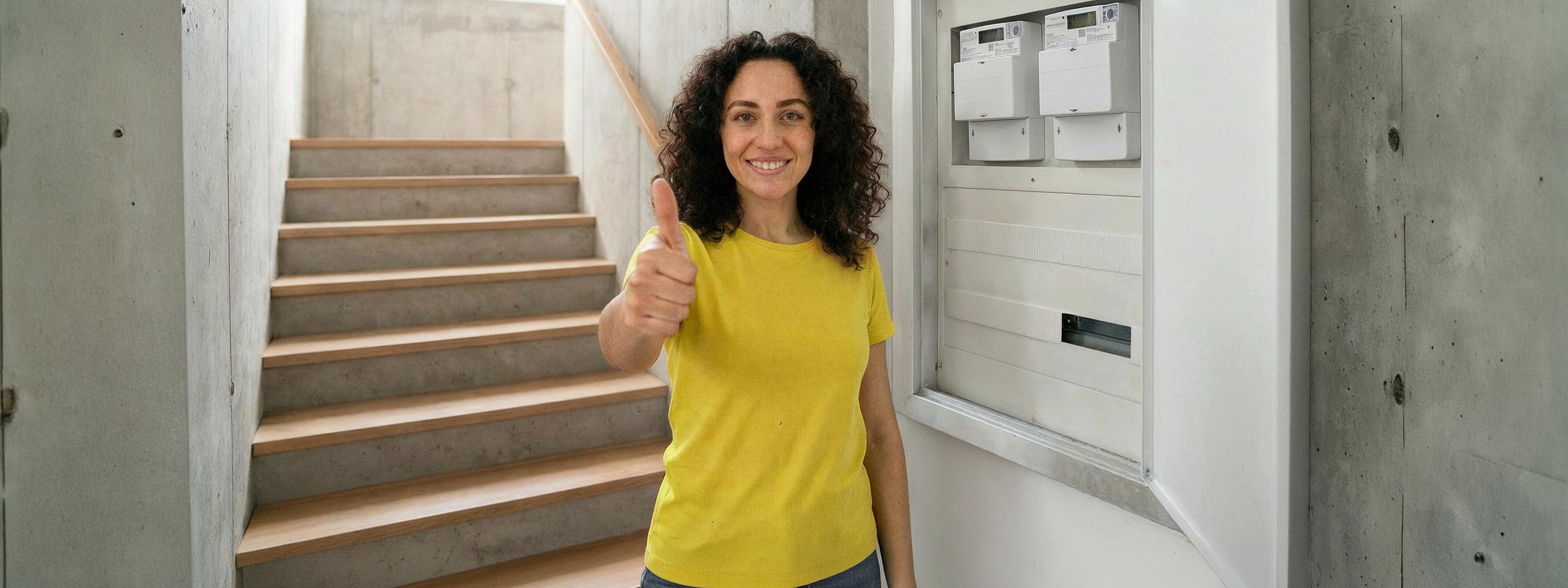 Eine Frau in gelbem Shirt steht neben Zählerkasten mit digitalem Stromzähler und zeigt lächelnd ihren Daumen hoch.