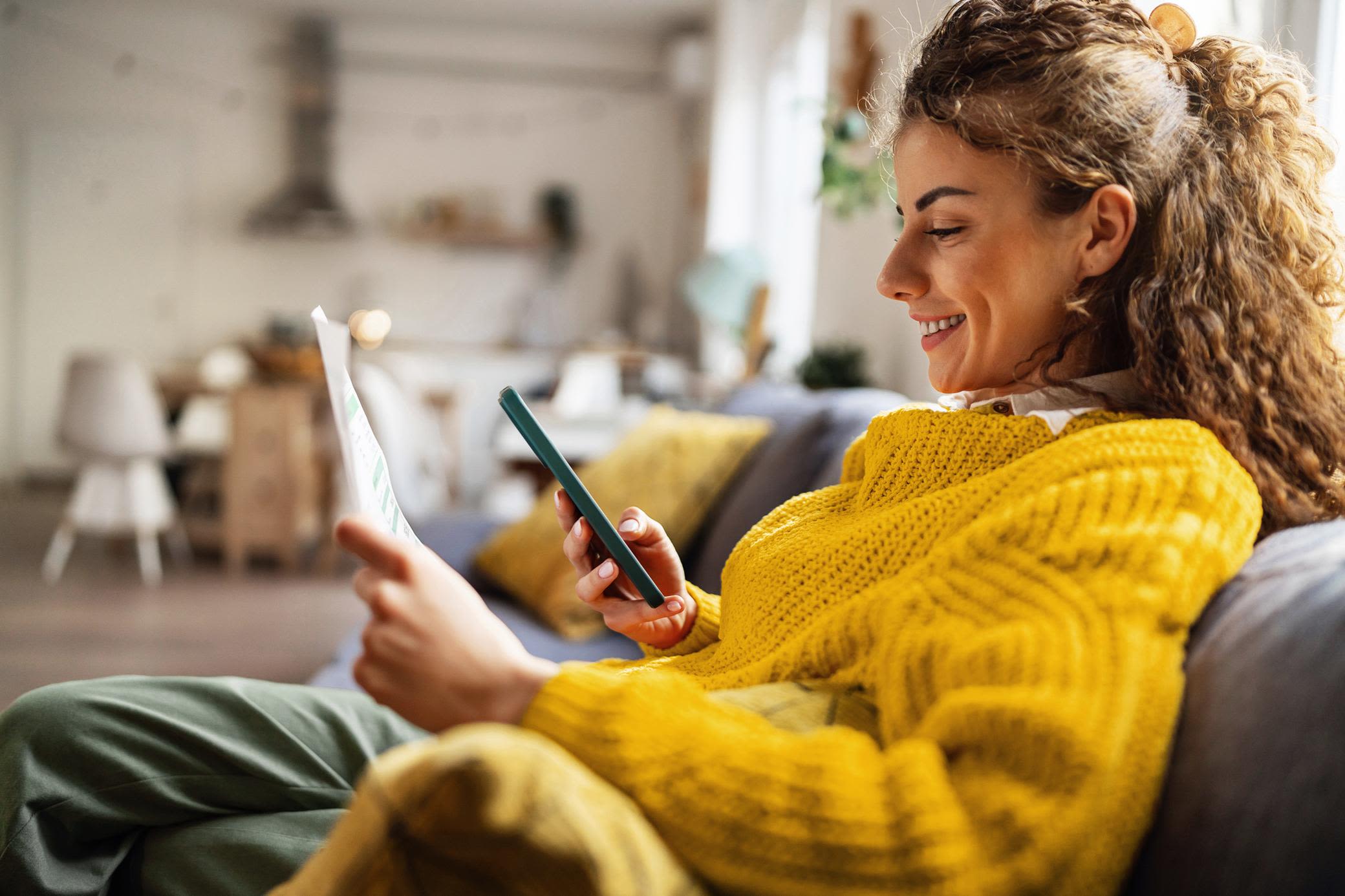 Deinen Strom einfach verstehen? Mit Yello geht das. Eine Frau sitzt auf dem Sofa und hält in der einen Hand einen Zettel, in der anderen ein Smartphone.
