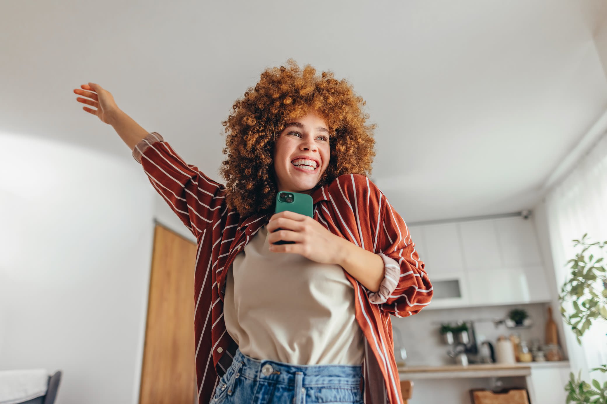 Die Zukunft der Energie ist grün - und du bist mit Yello ein Teil davon Fröhliche Frau mit grünem Handy streckt den Arm nach oben - eine Küche ist im Hintergrund