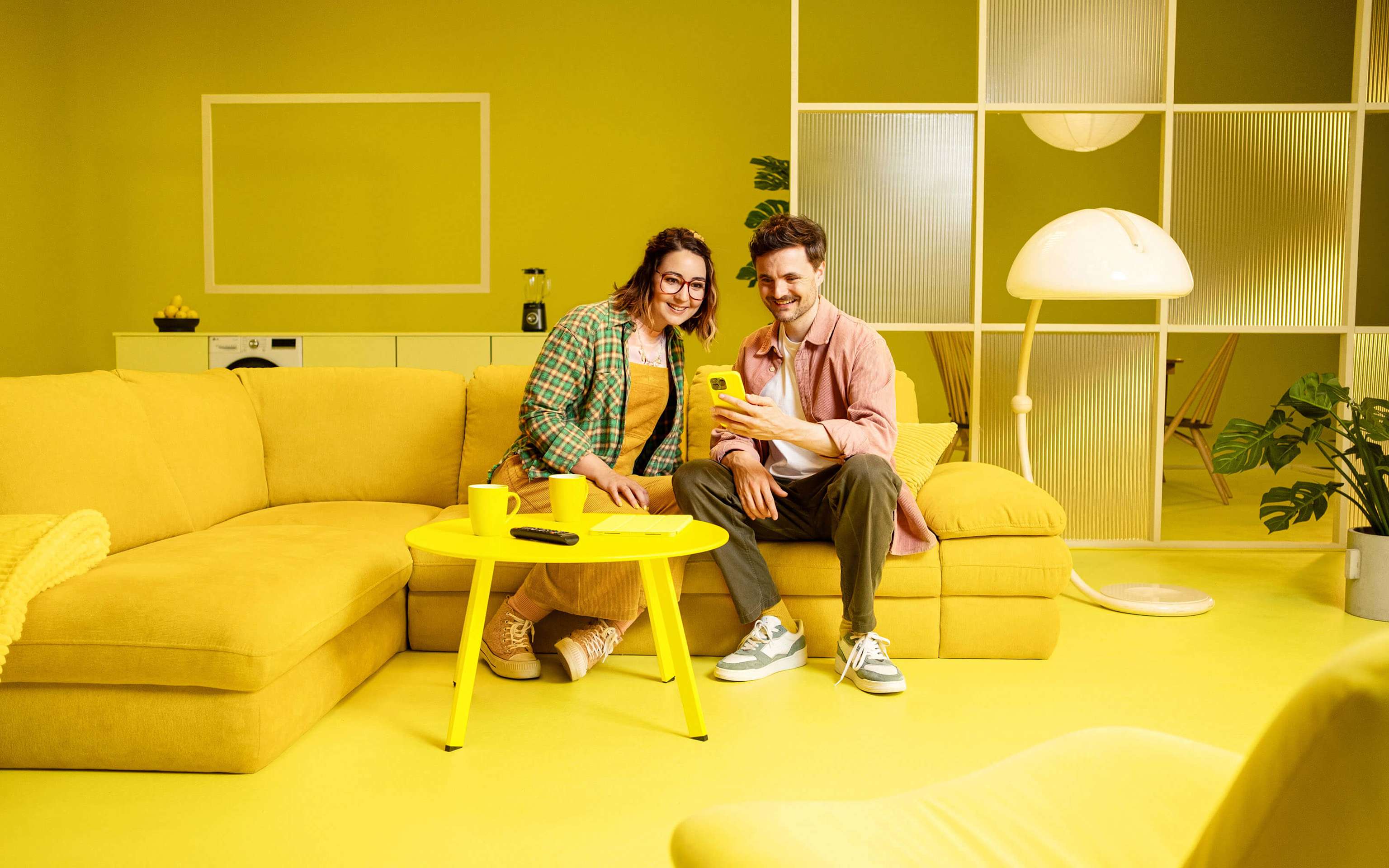 Ein junges Paars sitzt auf einer gelben Couch und sie schauen auf ein Smartphone mit der Yello App Ein junges Paars sitzt auf einer gelben Couch und sie schauen auf ein Smartphone mit der Yello App