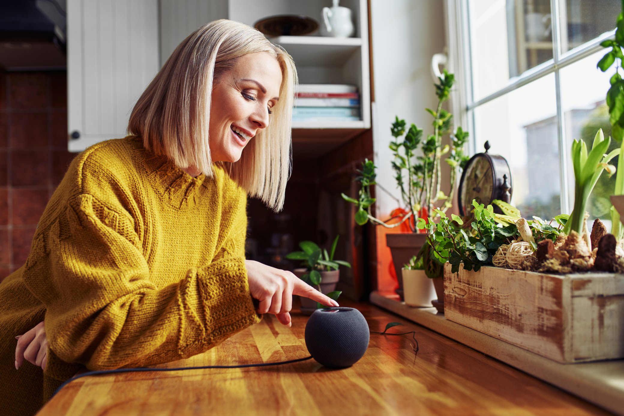 Smart Home Systeme machen dir den Alltag leichter Frau in gelbem Pullover tippt auf einen Smart Speaker, in einer Küche mit Pflanzen vor dem Fenster