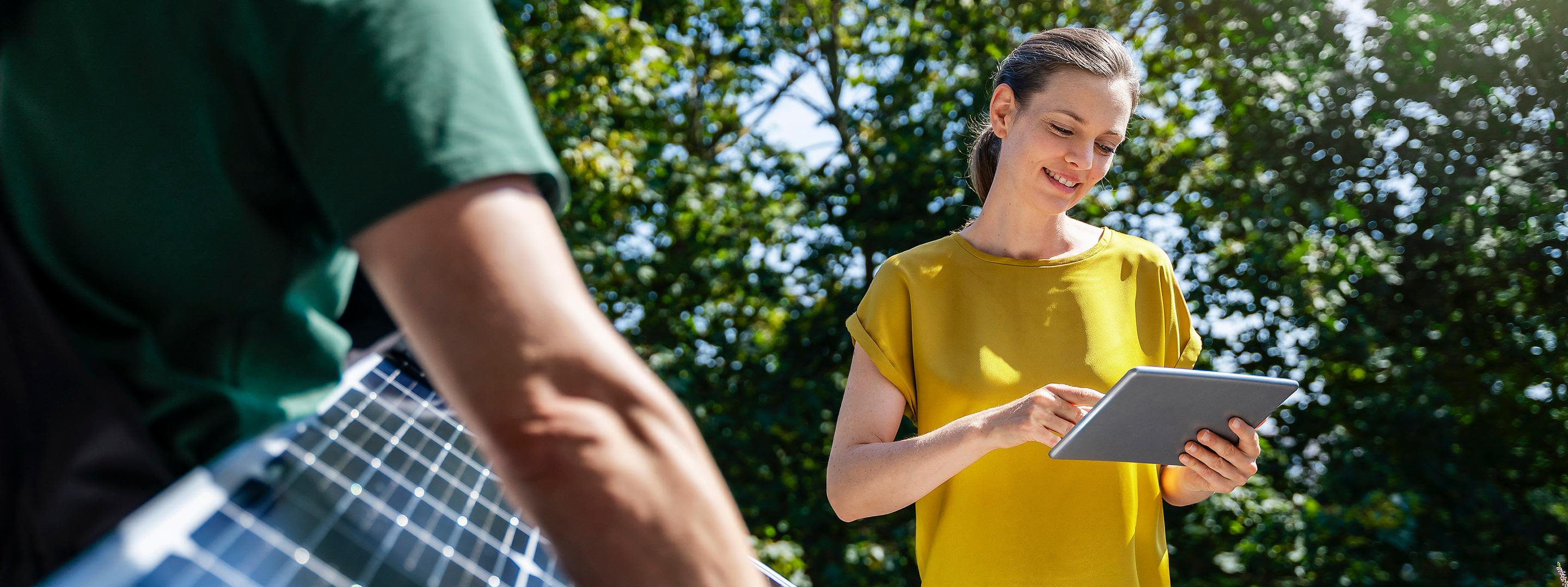 Mit Einspeisevergütung mehr Profitabilität für deine Solaranlage Frau im gelben Shirt steht in der Natur vor grünen Bäumen mit Tablet in der Hand und Mann mit grünem Shirt trägt ein Solarpanel
