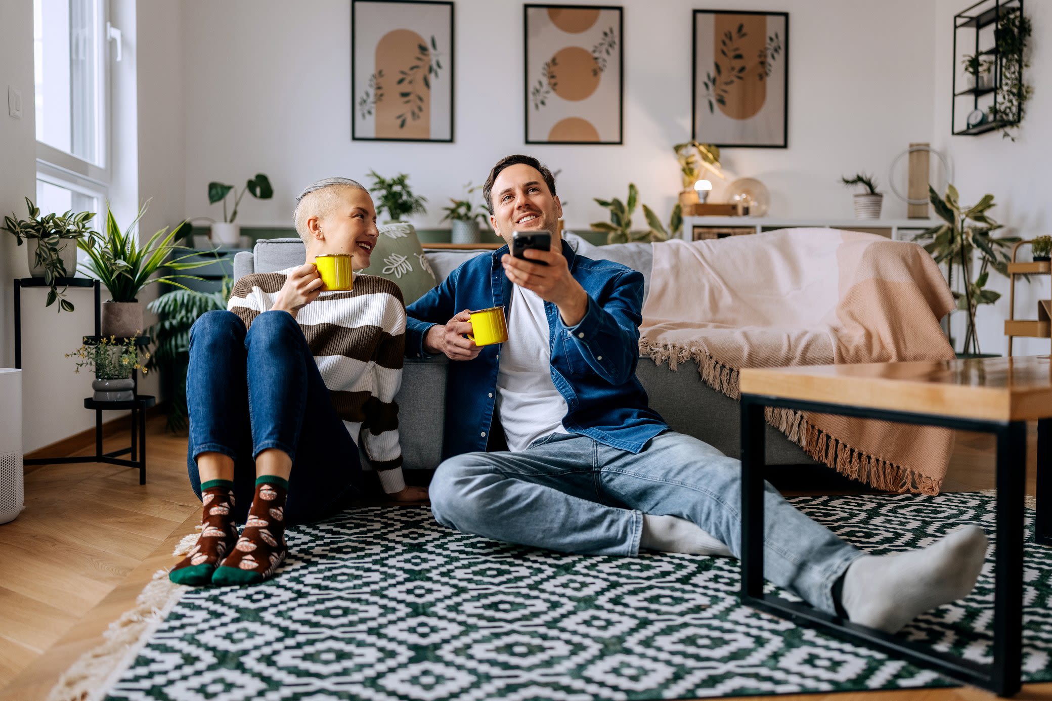 Digital und easy - Mit Smart Home optimierst du deinen Energieverbrauch Ein junges Paar sitzt mit einer Tasse Tee in der Hand auf dem Boden und steuert ein Smart-Home-System mit dem Smartphone
