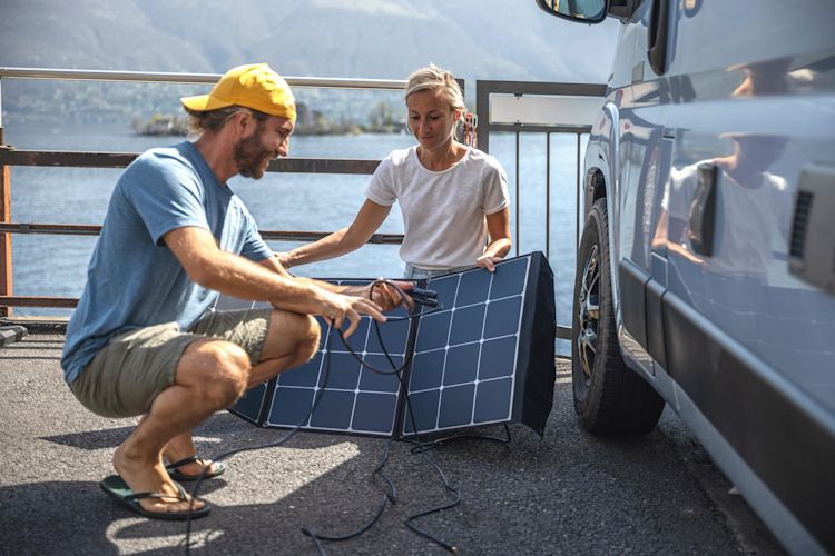 Mit Solar lädst du sogar unterwegs beim Camping deine Geräte Junges Paar mit faltbarem Solarpanel neben ihrem Camper Van