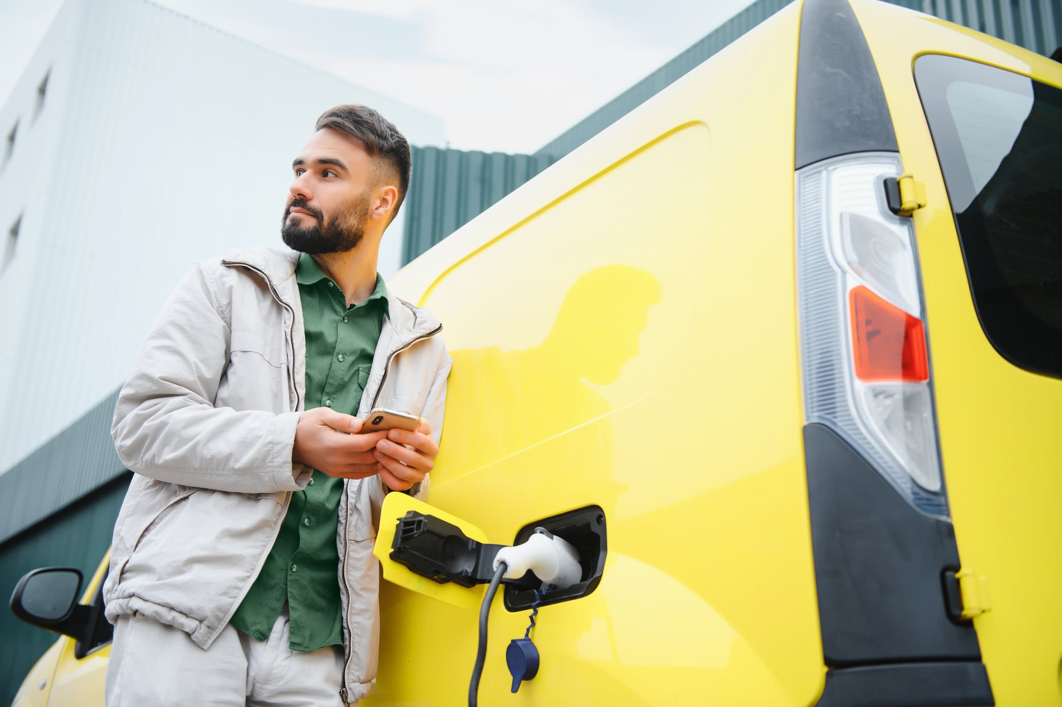 Für dein elektrisches Firmenfahrzeug bekommst du steuerliche Vergünstigungen Ein Mann mit Handy in der Hand lädt seinen elektrischen Minivan