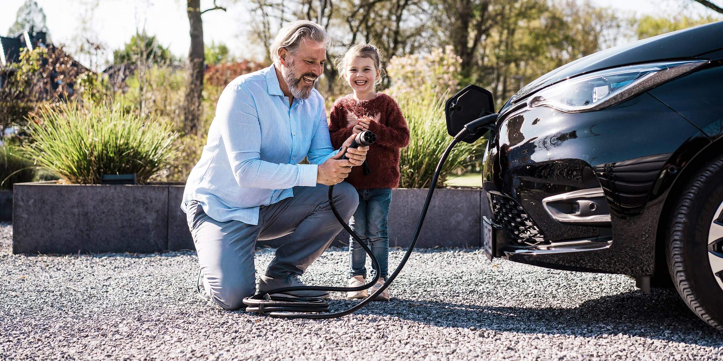 Mann mit Tochter, der an einem sonnigen Tag vor einem E-Auto einen Ladekabel in der Hand hält