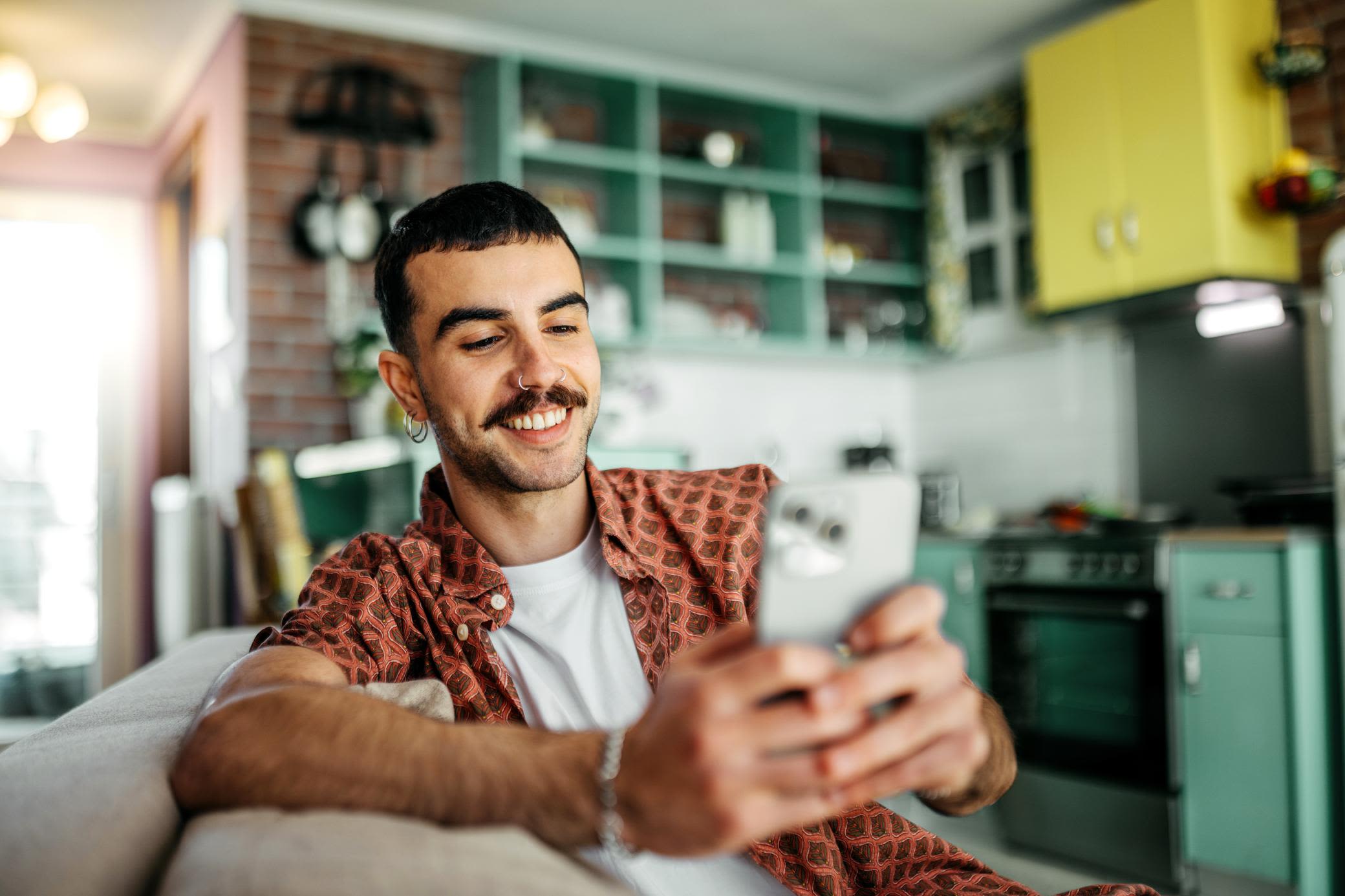 Mit App hast du deinen Energieverbrauch immer im Blick Ein Mann mit Handy sitzt auf der Couch und im Hintergrund ist eine grüne Küche