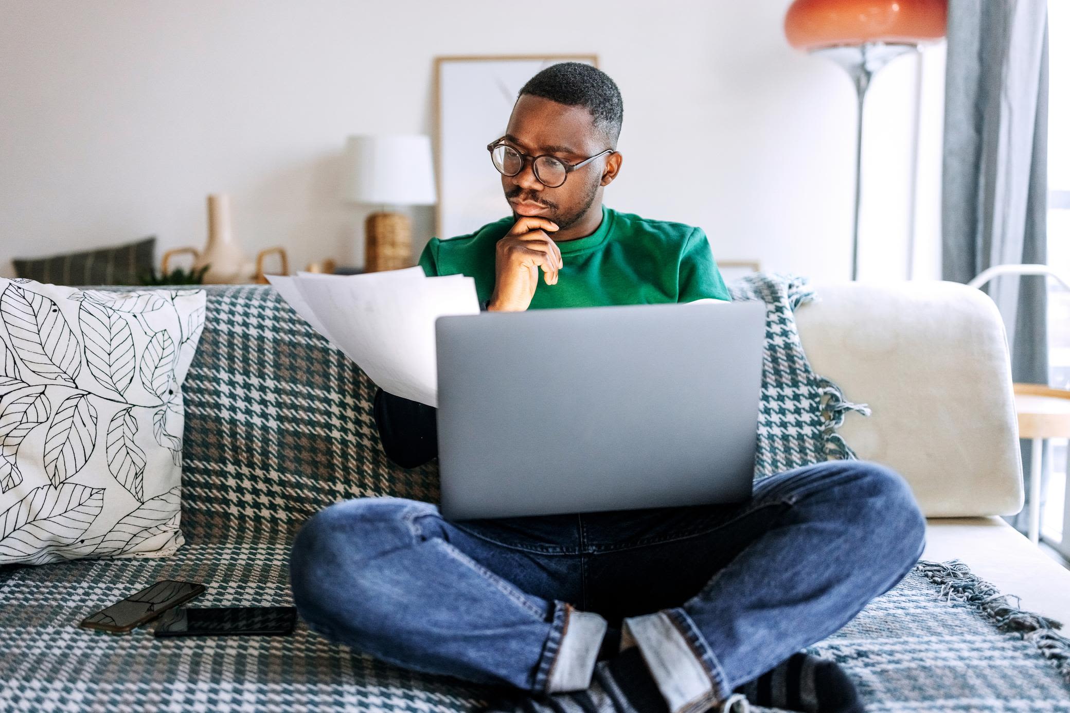 Energie wird digital und damit viel transparenter Junger Mann sitzt auf Sofa mit Laptop wie Papieren und überlegt
