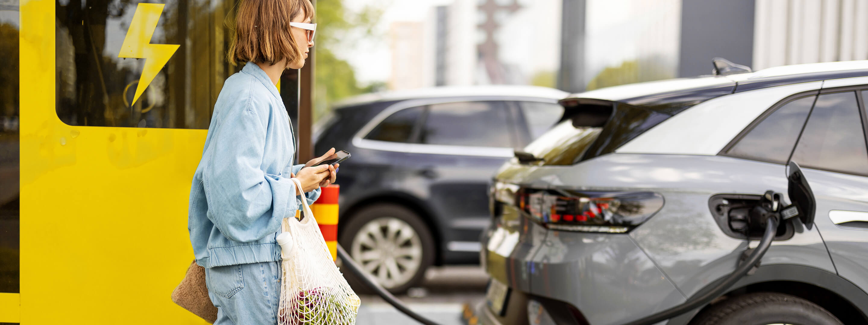 Frau mit Handy und Lebensmitteln wartet auf Laden des Elektroautos 