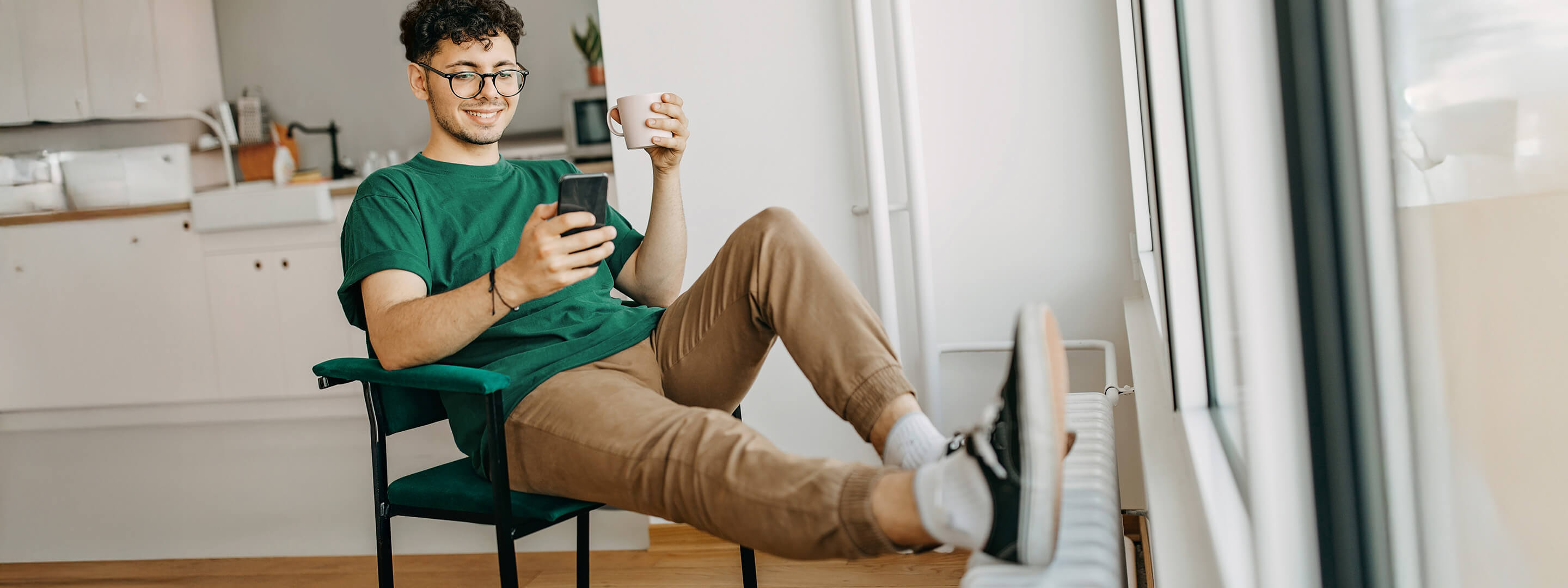 Junger Mann sitzt lächelnd am Handy, mit den Füßen auf der Heizung