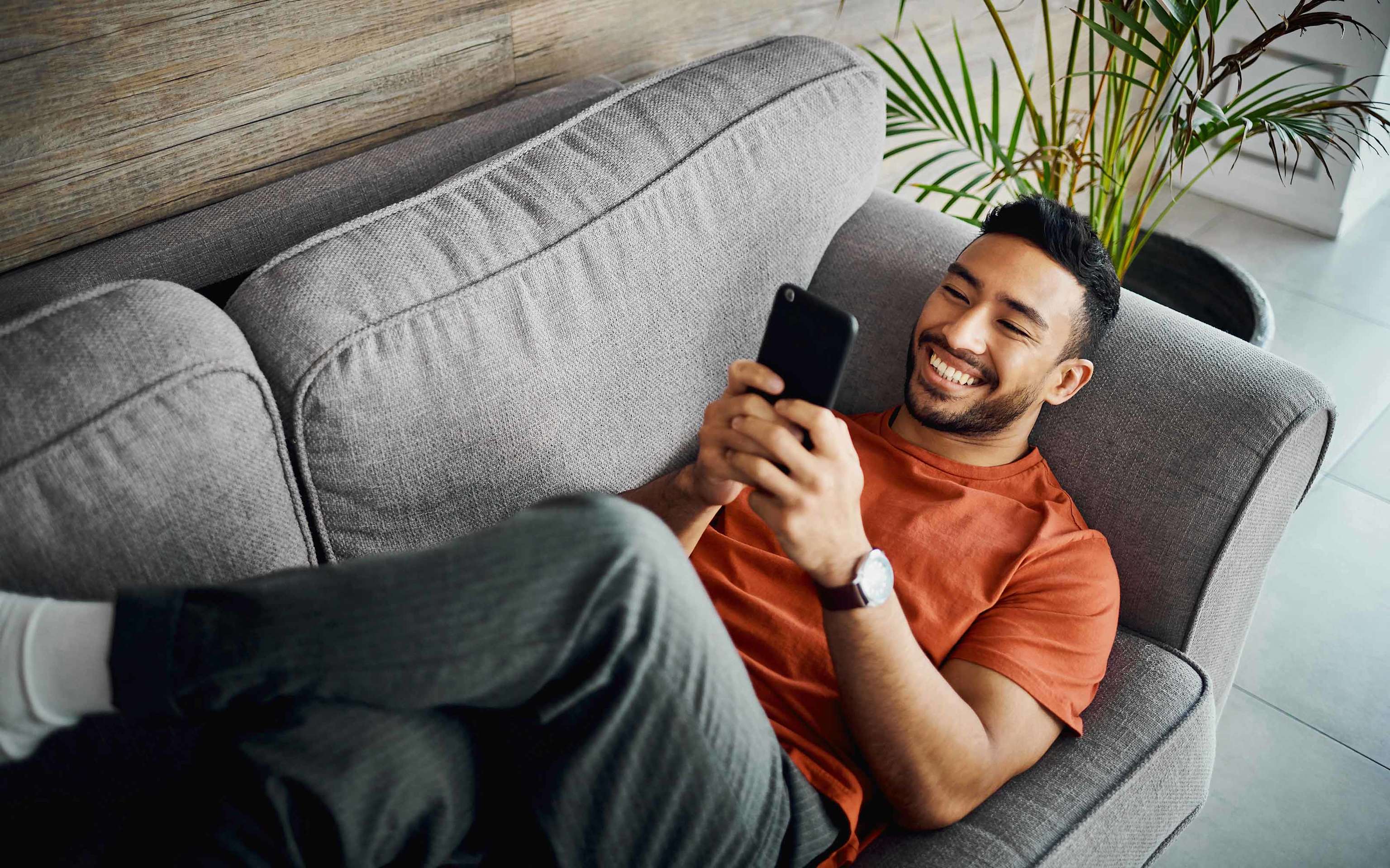 Junger Mann liegt lächelnd auf dem Sofa und schaut entspannt auf sein Smartphone Junger Mann liegt lächelnd auf dem Sofa und schaut entspannt auf sein Smartphone