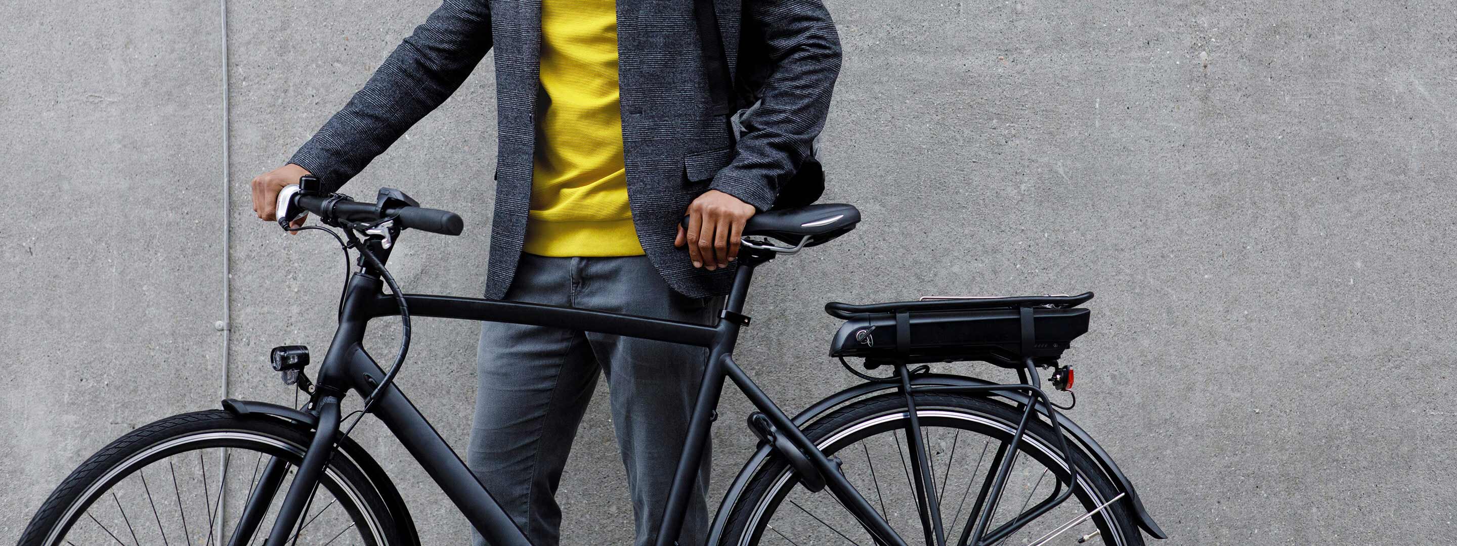 E-Bike günstig kaufen. Ob für City- oder Bergtouren. Mann steht mit seinem E-Bike Fahrrad vor einer grauen Wand