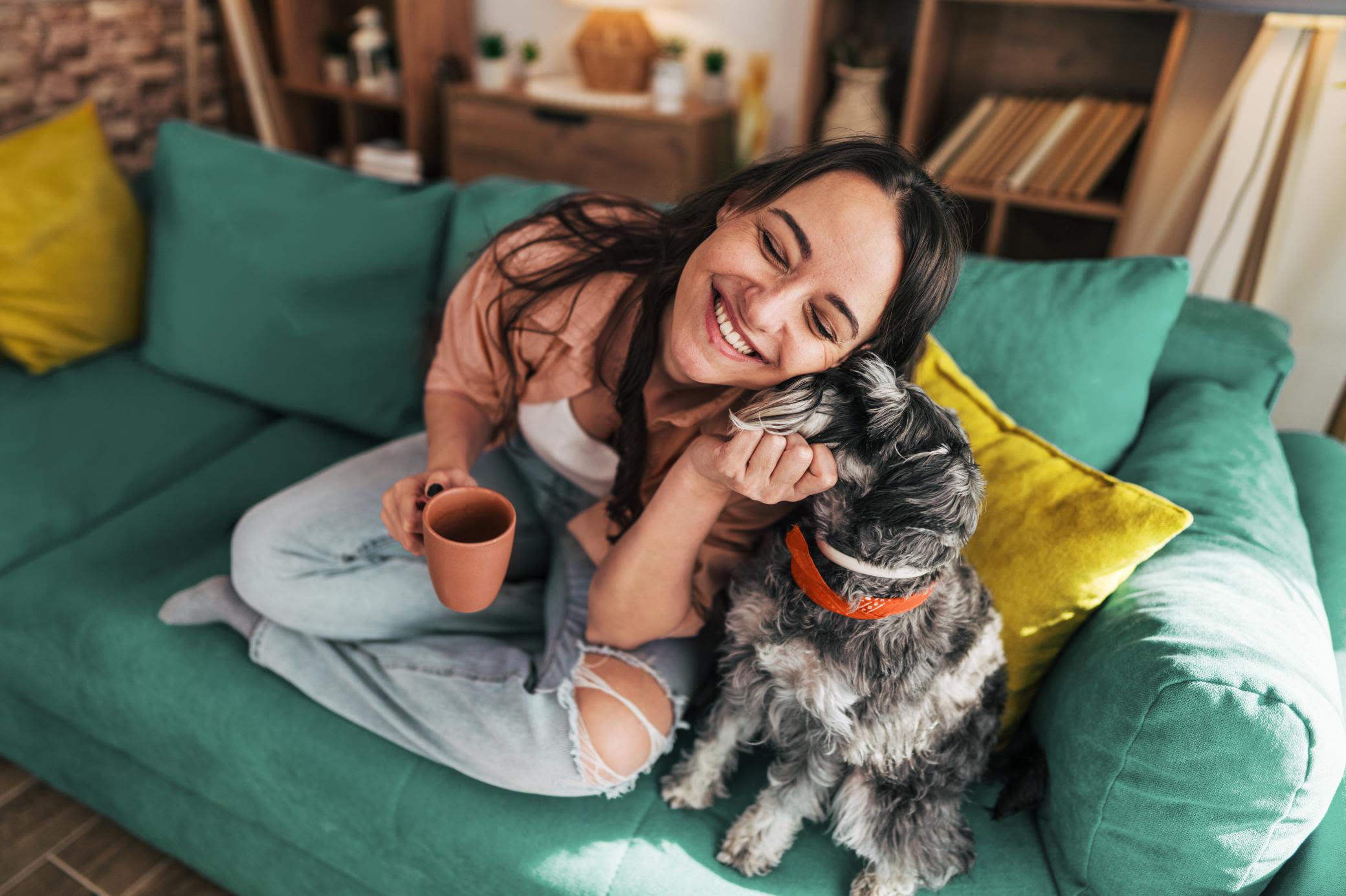 Feeling good - mit guter Energie und guten Informationen Eine junge Frau lächelt, während sie eine Tasse Kaffee trinkt und mit ihrem Hund auf dem Sofa sitzt