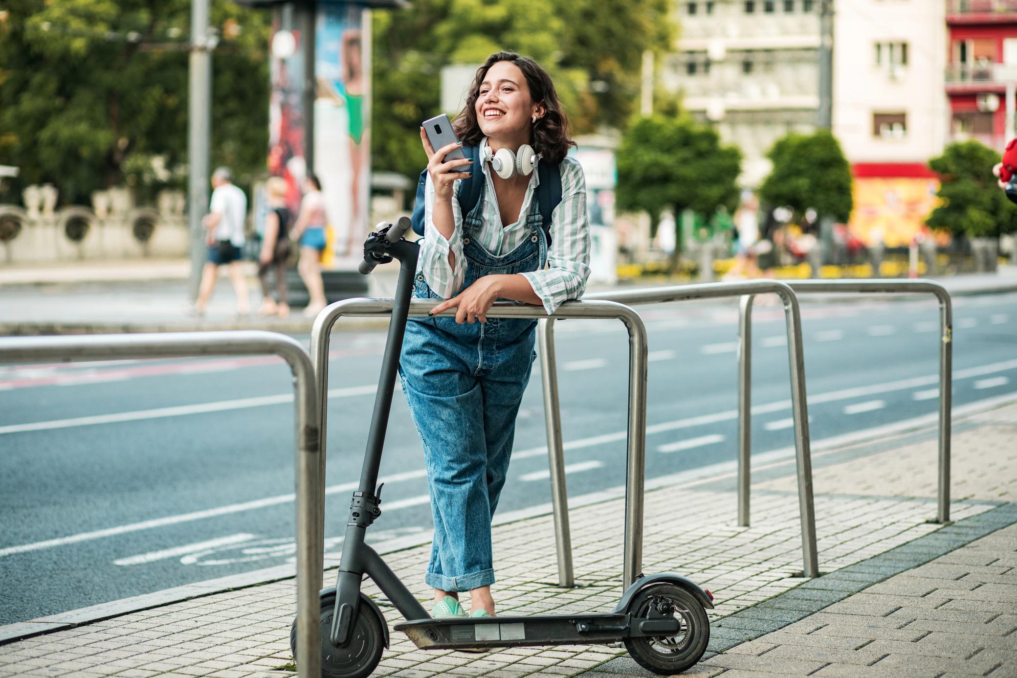 Frau mit Smartphone in der Stadt lehnt neben ihrem E-Scooter