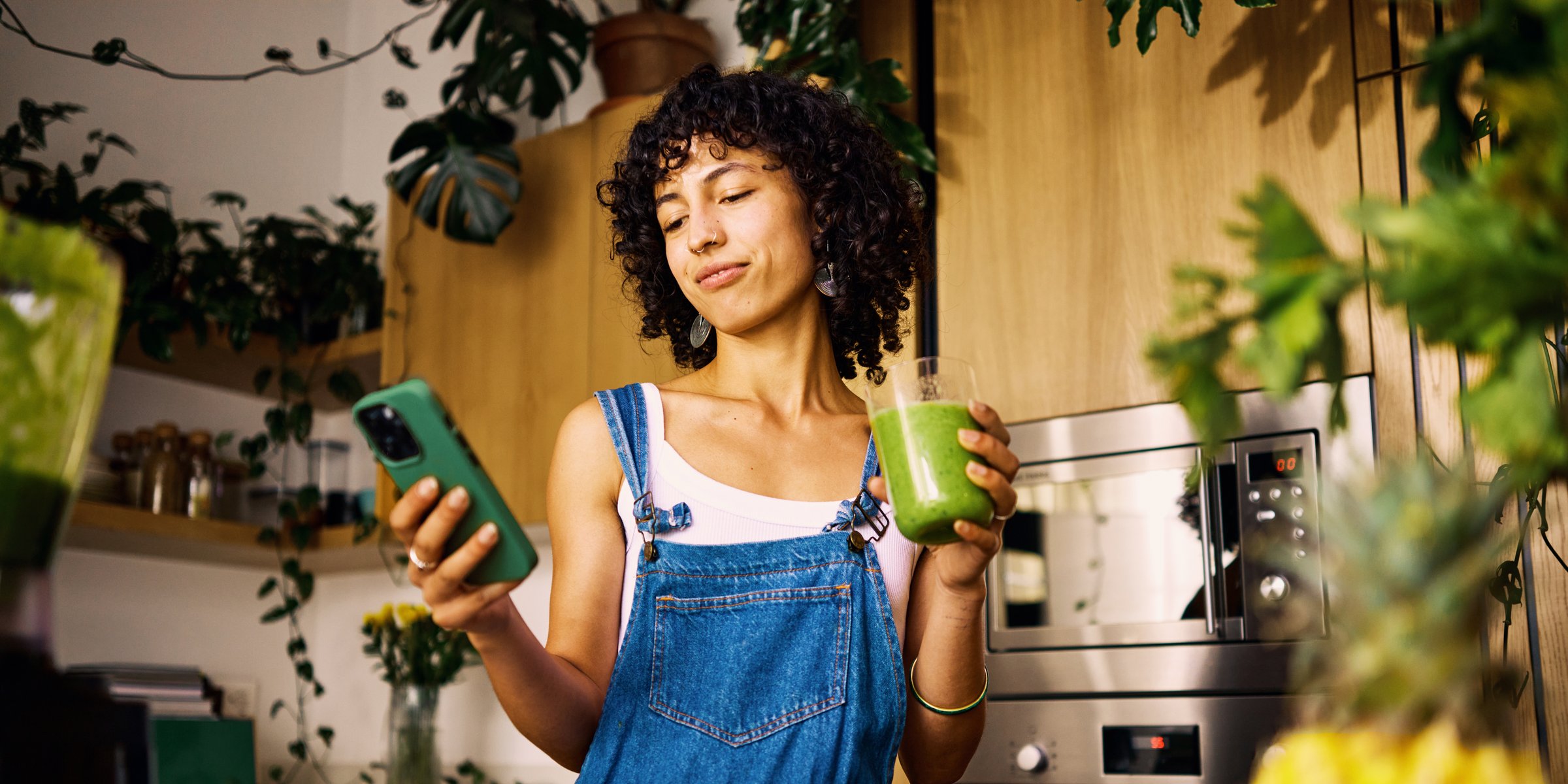 Frau mit Smoothie in der Hand steht in der Küche und schaut auf ihr Smartphone