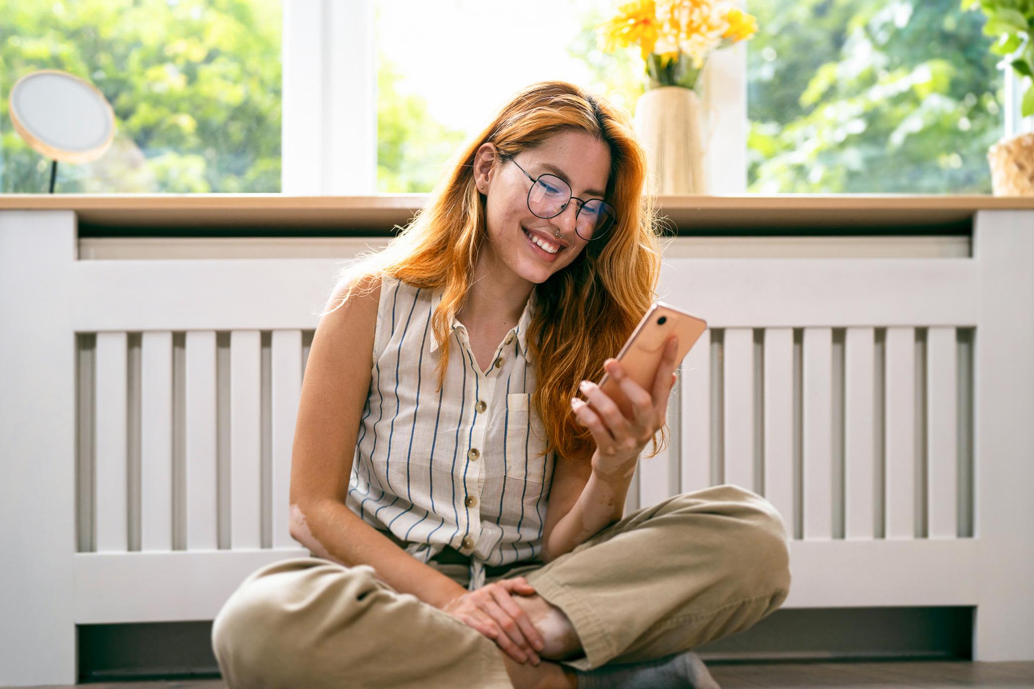 Beim Heizen sparen: mit grüner Energie und passenden Tarifen Lächende Frau sitzt auf dem Boden vor der Heizung und schaut aufs Handy