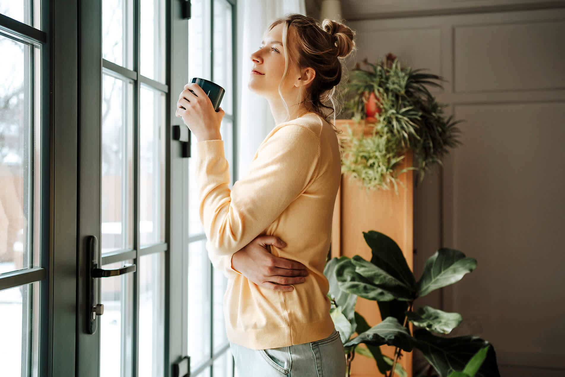 Eine junge Frau steht mit einem Kaffeebecher am Fenster in ihrer Wohnung und schaut hinaus Eine junge Frau steht mit einem Kaffeebecher am Fenster in ihrer Wohnung und schaut hinaus