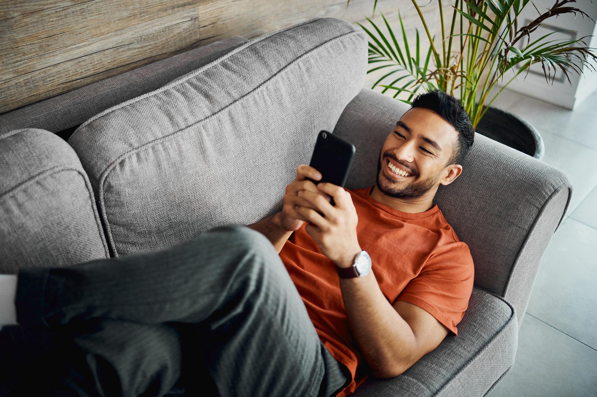 Appsolutely digital - so regelst du heute dein HEMS bequem vom Sofa aus Junger Mann mit orangenem Shirt liegt mit überkreuzten Beinen auf dem Sofa und schaut mit Freude aufs Handy