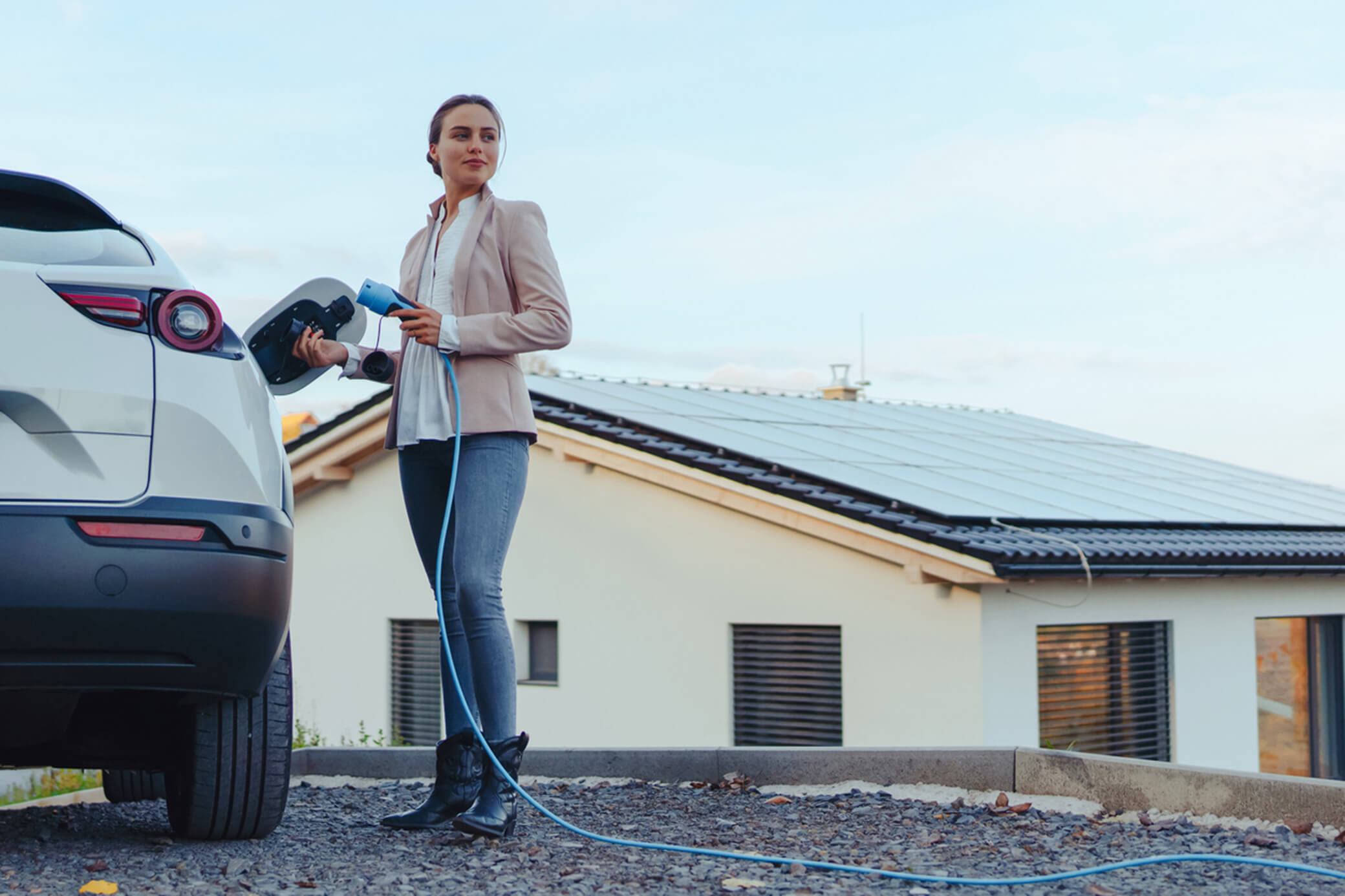 Mit Sonnenstrom lädst du dein E-Auto noch nachhaltiger Eine Frau lädt ein weißes Elektroauto und hält ein Ladekabel in der Hand - im Hintergrund ist eine Solaranlage
