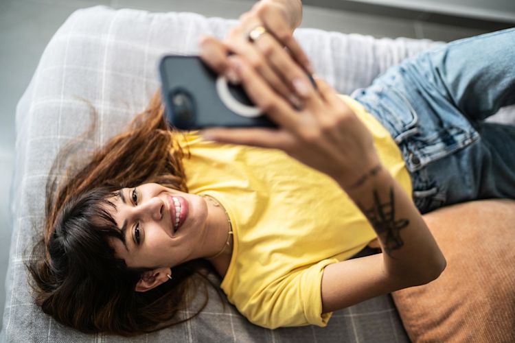 Wechsel deinen Energieanbieter entspannt Online Eine junge Frau im gelben T-Shirt liegt auf dem Sofa und schaut aufs Handy