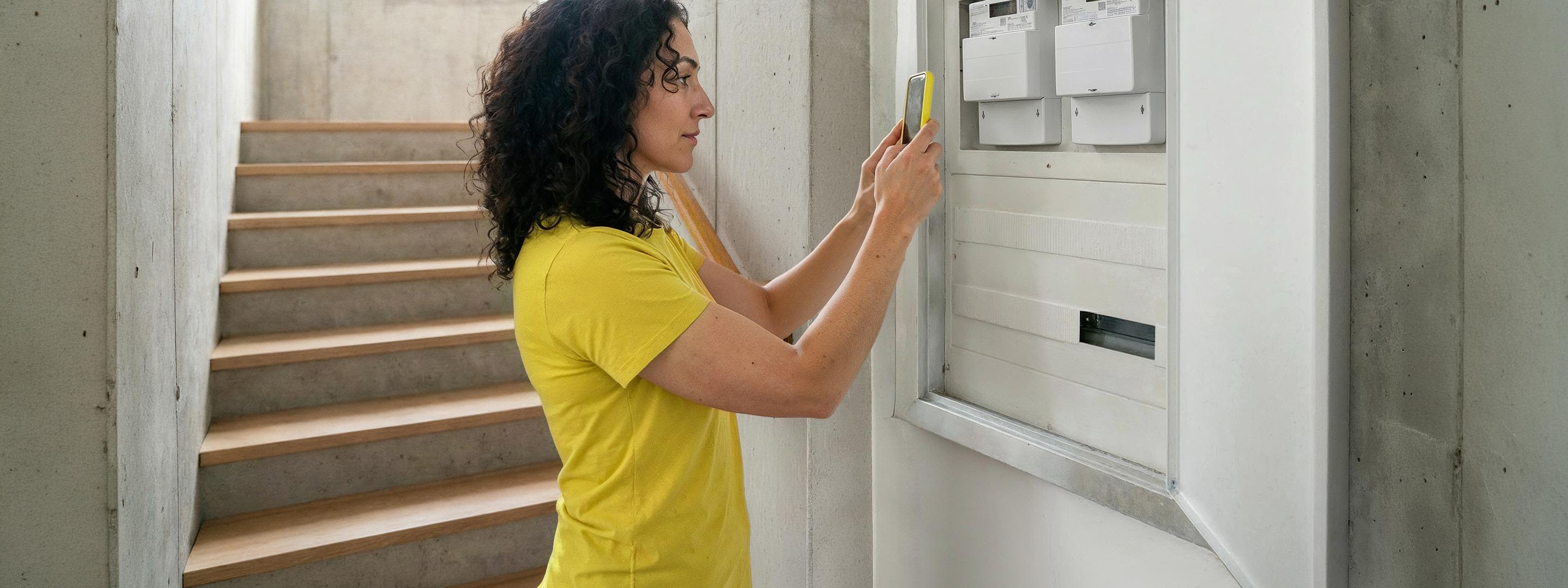 Stromzähler ablesen: So funktioniert's einfach immer Frau mit braunen Locken und gelbem Shirt steht vor Zählerkasten und fotografiert Zählerstand mit gelbem Handy und der Yello App.