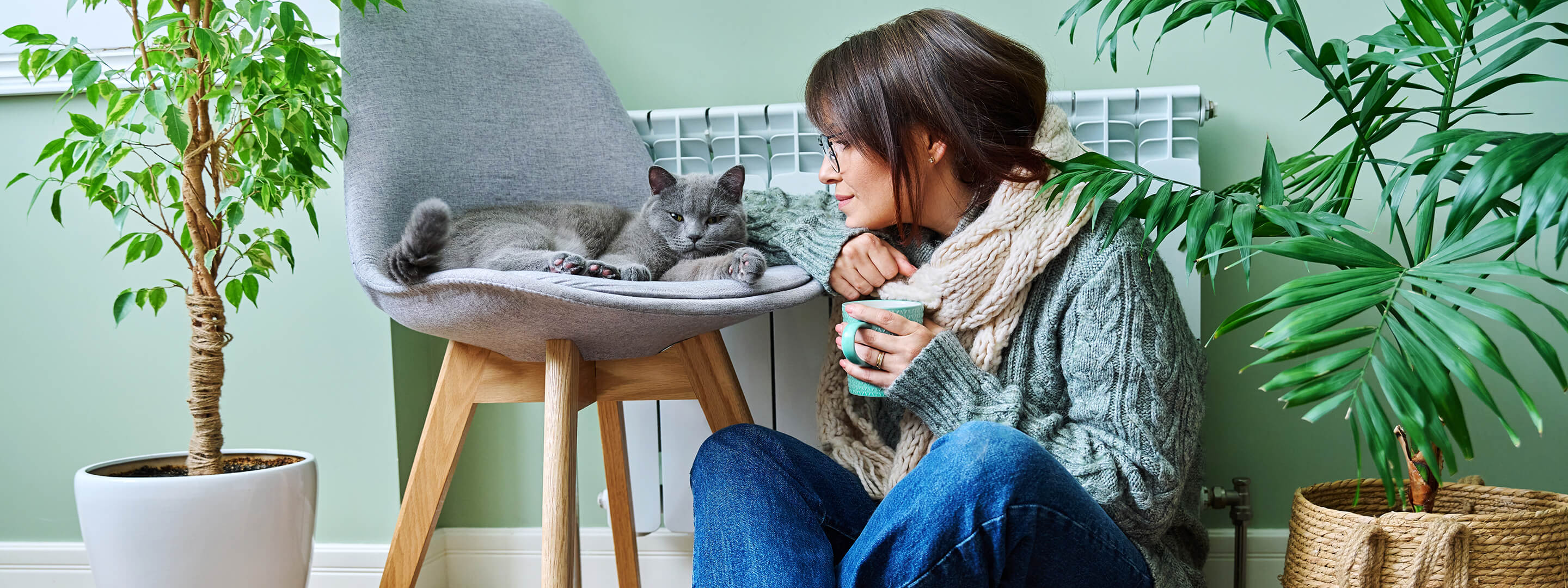Mit Heizstrom wohlig warm. Günstiger mit Strom heizen Frau sitzt in warmer Kleidung mit Katze vor der Heizung