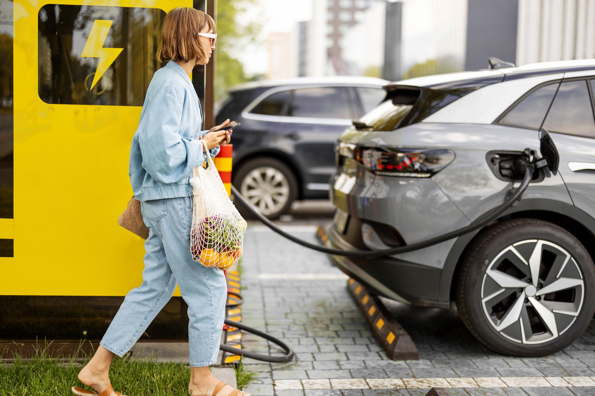 E-Auto schnell in der Stadt oder Zuhause laden Frau mit Handy und Lebensmitteln wartet auf das Laden ihres Elektroautos in der City