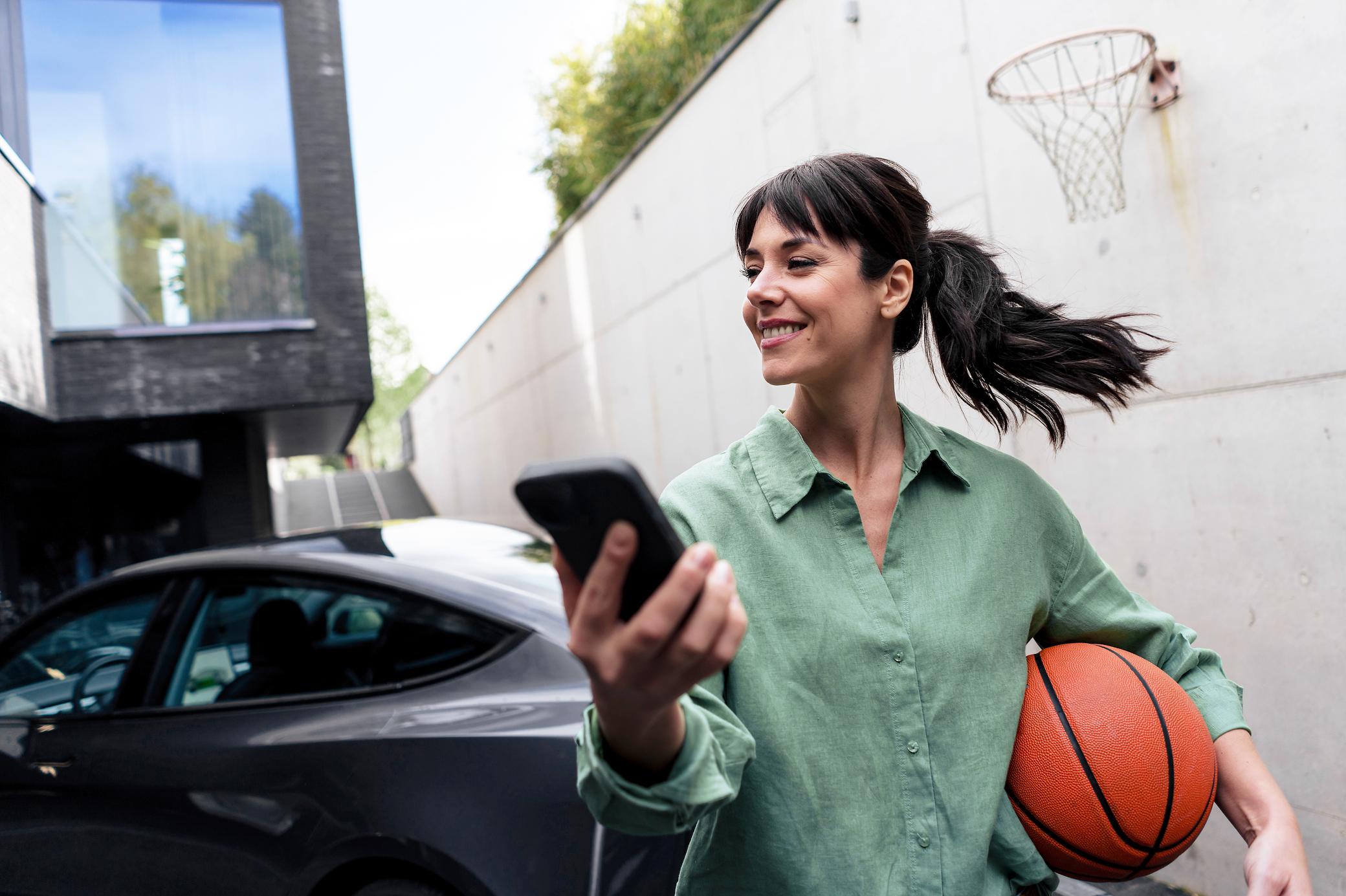 Lachende Frau mit Basketball unter dem Arm und Handy in der Hand lädt ihr E-Auto smart zuhause