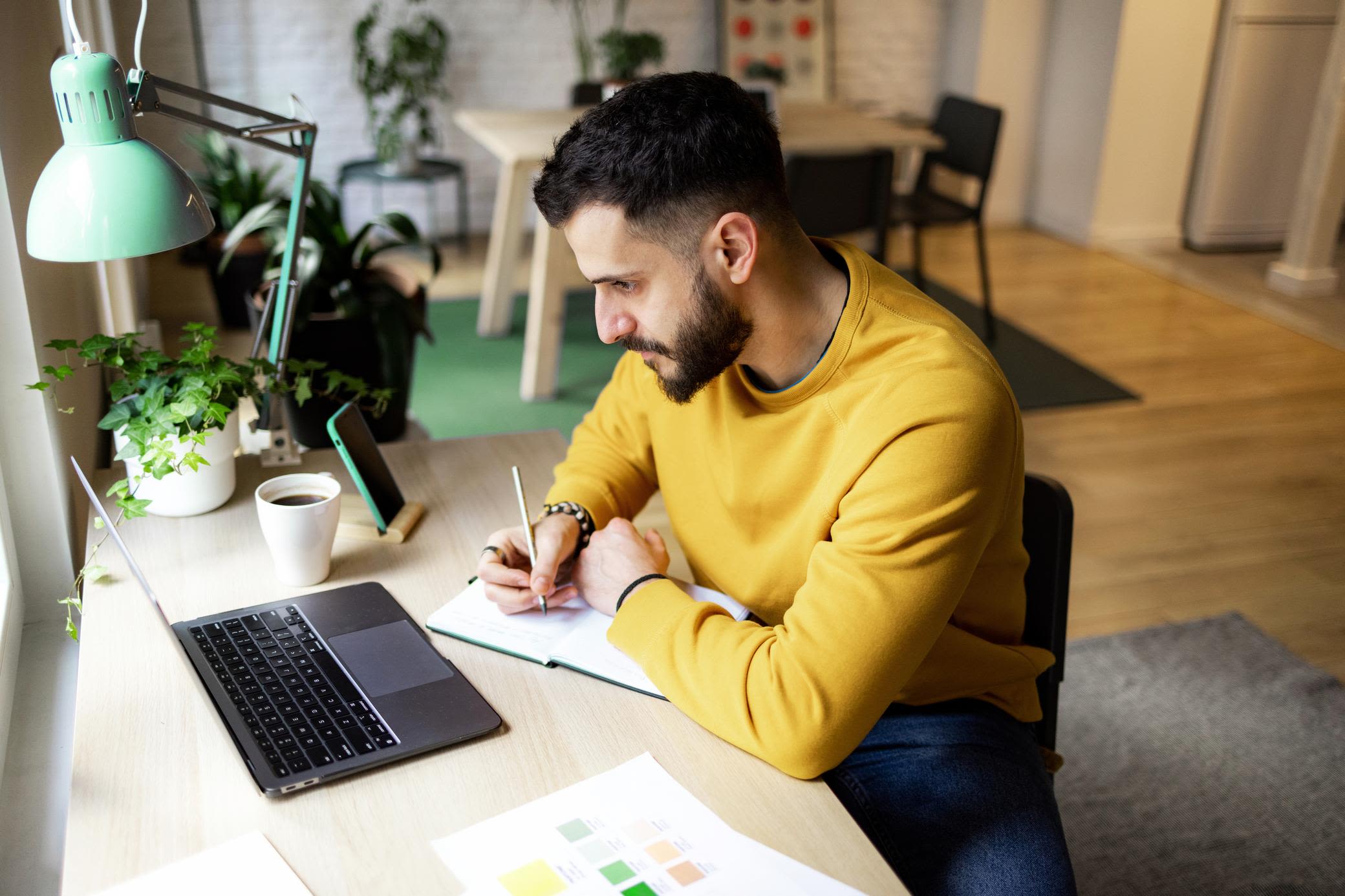 Bleibt gut informiert über Energie und Nachhaltigkeit Junger Mann sitzt zuhause am Laptop am Schreibtisch schreibt in sein Notizbuch