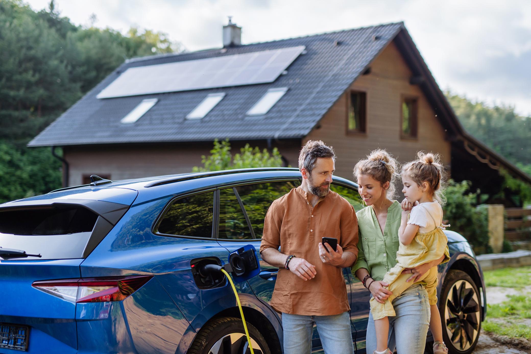 Familie mit Mädchen laden Elektroauto vor dem Haus mit Solaranlage auf dem Dach