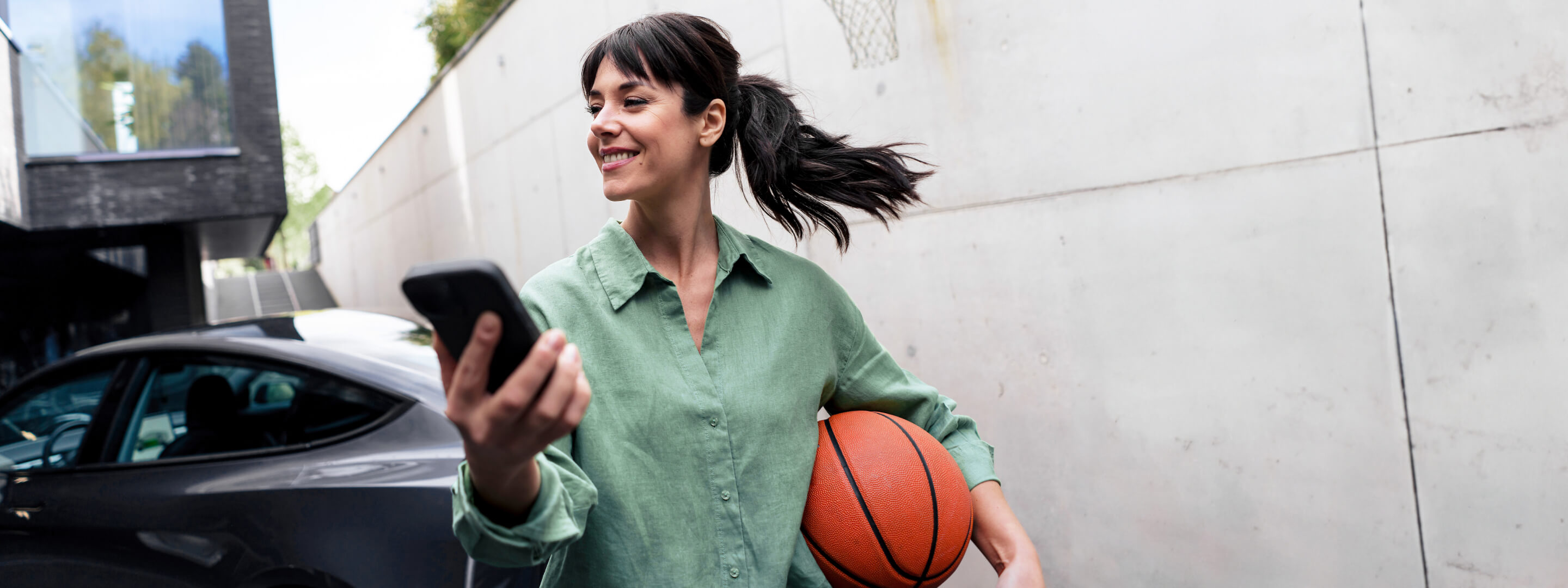Frau mit Basketball und Handy lädt ihr E-Auto zuhause