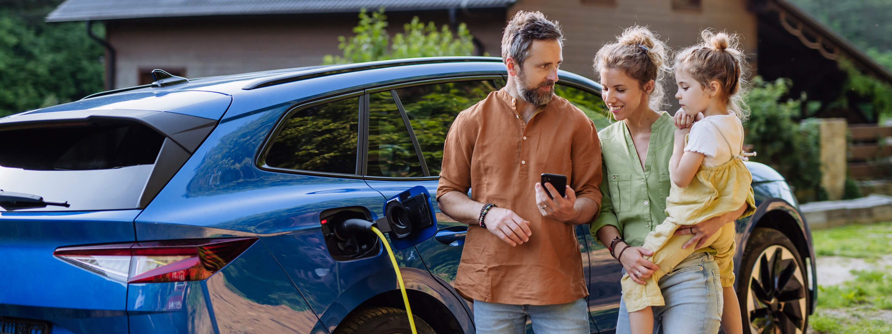 Bei Kauf eines Elektroautos gibt es einiges zu beachten. So geht's einfach. Familie, bestehend aus Vater, Mutter und Tochter, steht vor einem blauen Elektroauto, das gerade geladen wird. Alle blicken auf das Handy des Vaters.