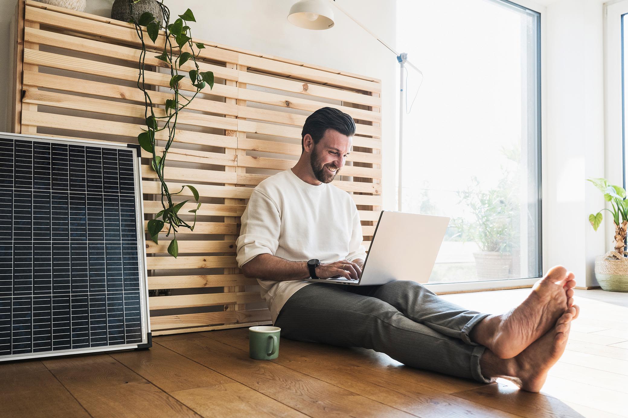 Balkonsolar für zuhause ist einfach und günstig Mann der neben einem Solarmodul sitzt und einen Laptop benutzt