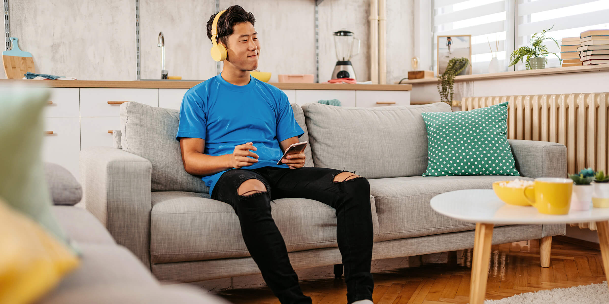 Junger Mann im blauen T-Shirt und gelben Kopfhörern sitzt auf der Couch und hört Musik